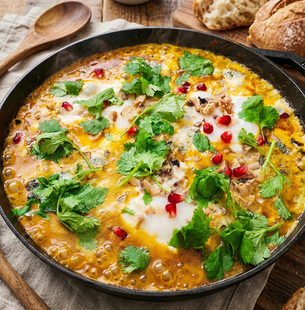 Herzhaftes Kürbis-Shakshuka in einer schwarzen Pfanne, garniert mit Eiern, Maronen, Roquefort, Granatapfelkernen und Koriander.